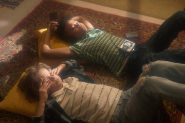 Renkli halıda uzanan iki çocuğun yüksek açılı görüntüsü, birinin yüzünde elleri, diğerinde klasik müzik çalar var.