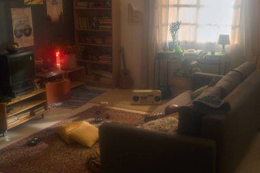 Televizyon, pikabın, teybin, gitarın, kitaplığın ve pencereden süzülen güneş ışığının da dahil olduğu klasik dekorlu rahat bir oturma odası.