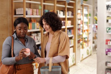 İki genç bayan müşteri yeni yüz kremini tartışırken Afrikalı Amerikalı kadın plastik kavanoz hakkında bilgi veriyor.
