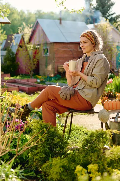 stock image Serene female gardener or farmer in workwear sitting on chair among green plants and flowers and having cup of tea or coffee
