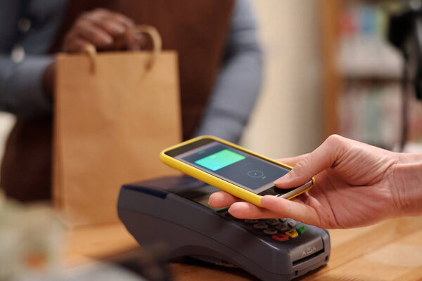 Hand of young female shopper with smartphone making contactless transaction while holding gadget over payment terminal