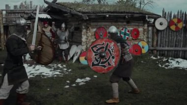 İki genç erkek ortaçağ savaşçısının miğferleri, kalkanları ve kılıçlarıyla ahşap kalenin avlusunda antrenman yaparken, arkadaşları izlerken ve tezahürat yaparken tam çekimi.