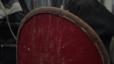 Zincirli kasklı, kırmızı kalkanlı, kılıçlı, tahta kapının yanında duran, okçu yayı, oku hazırlayan, düşman saldırısına hazırlanan genç, beyaz sakallı savaşçının yakın çekim görüntüsü.