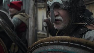 Miğfer ve zırh giymiş, elinde kılıç ve kalkanla kalenin kapısında yoldaşlarıyla düşman saldırısına hazırlanan olgun bir erkek ortaçağ Avrupalı askerinin gergin yüzünü yakından çek.