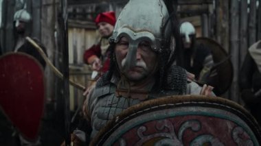 Zırhlı göğüs zırhı, miğfer, kalkan ve kılıç kuşanmış, feodal kalenin silahlı muhafızlarıyla kameraya bakarken çekilmiş orta boy bir portre resmi.
