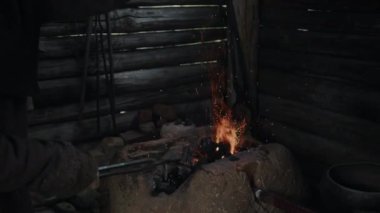 Orta yaşlı, siyah kapüşonlu demircinin el yapımı metal maşayla demir ocağında körükle kömür üflerken orta ölçekli bir fotoğrafı.