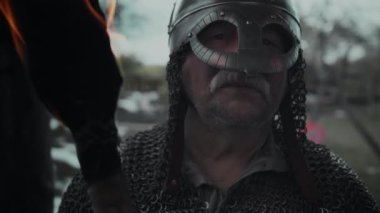 Zincirli tunik ve miğferli, gri sakallı, akşamları dışarıda devriye gezen, elinde meşale tutan ve uzaktan bakan olgun beyaz erkek Orta Çağ savaşçısının yakın plan portresi.