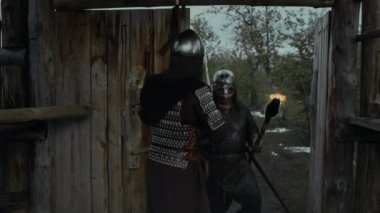 Zırhlı tunik ve miğferli feodal kale muhafızlarının devriyeden dönen, yürüyen, kılıç taşıyan ve yanan meşaleleri taşıyan yoldaşları içeri almak için ahşap kapıyı açtığı orta boy tam bir izleme görüntüsü.