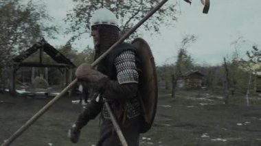 Zırhlı tunik giymiş, miğferli, sırtında boyalı kalkan, omzunda mızrak taşıyan, ahşap garnizonda yürüyen Orta Çağ erkek askerinin orta boy görüntüsü.