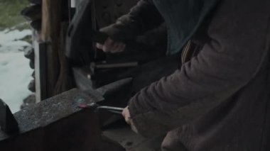 Kapüşonlu pelerinli ve köylü paltolu genç erkek demircinin elinde kızgın demir maşayla çekiçle örse de demircilikte ağır işçilik yaparken orta boy yakın çekimde çekilmiş bir fotoğrafı.