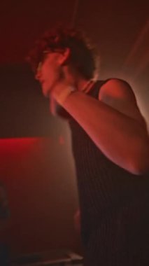 Kolsuz tişörtlü ve tekno çılgın güneş gözlüklü genç adamın el kamerasıyla çekilmiş dikey görüntüsü kırmızı neon ışıklı gece kulübü ile elektronik dans müziğine güçlü hareketler yapıyor.