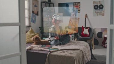 Yan çekim, genç bir kızın günlük kıyafetleriyle yatağında bacak bacak bacak üstüne atarak meditasyon yapması AR kulaklığı takması ve huzurlu ve sakin gün batımı manzarasına bakması.