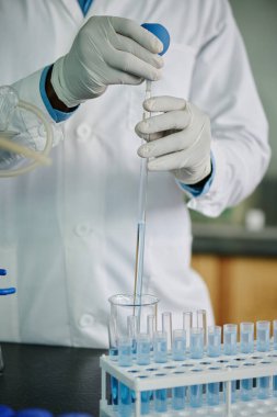 Modern kimyagerin eldivenli elleri ya da laboratuvar önlüklü araştırmacı deney sırasında cam borudan tüpe sıvı madde atıyor.