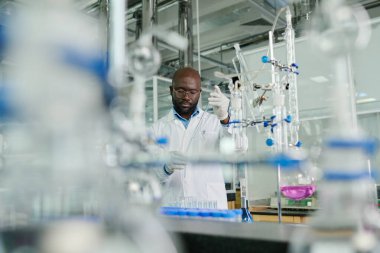 Laboratuvar önlüğü ve eldivenleriyle kimyasal araştırma yaparken cam eşyalar kullanan genç Afrikalı Amerikalı erkek araştırmacı.