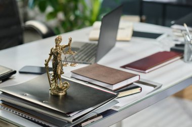 Small statuette of blindfolded goddess of Justice Themis with scale and sword in hands standing on stack of folders against other supplies clipart