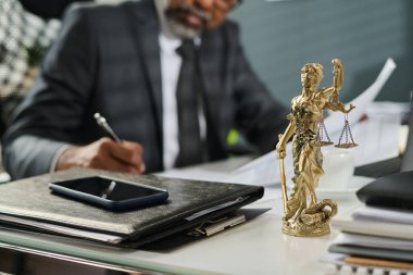 Statuette of Themis standing on workplace of lawyer with mobile phone on top of stack of folders containing juridical paper documents clipart