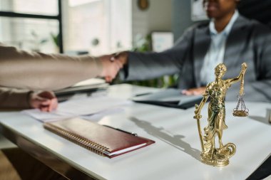 Small bronze statuette of Themis standing on workplace of lawyer with notepad in leather cover against two solicitors shaking hands clipart