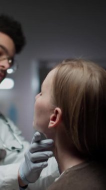 Dikey olarak genç sakallı çift taraflı doktor gözlük takıyor, önlük takıyor ve steteskop boynuna takıyor. Kadın hastayı muayene ediyor. Klinikte her yıl yapılan kontroller sırasında boynundaki gangliyonları muayene ediyor.