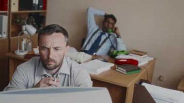 Bıyıklı, sakallı, klasik bilgisayar üzerinde çalışan, ofiste sigara içen, arka planda telefonla konuşan çeşitli patronların el kamerasıyla çekilmiş resmi.