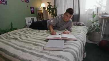 Genç işitme engelli beyaz erkek öğrencinin evde yatakta yatması, kulağına koklear implantı yerleştirmesi, kolej ödevi için ders kitabı okuması.