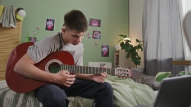 Orta boy işitme engelli, koklear implant takan, evde tek başına oturan, online ders ile akustik gitar çalmayı öğrenen beyaz bir çocuk.
