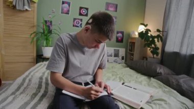 Evde ders kitaplarıyla yatakta oturan, defterine yazı yazan, kameraya bakıp gülümseyen, koklear implantı olan 16 yaşında, neşeli, sağır bir erkek öğrencinin orta boy portresi.