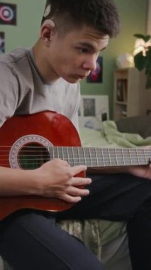 Kafasına koklear implant takmış işitme engelli bir gencin orta boy dikey pozu. Yatak odasında oturup online ders izlerken gitar çalmayı öğreniyor.