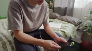 Beyaz tenli bir gencin evde yatağında oturmuş, koklear implantı akıllı telefona bağlayıp müzik dinlerken, ritme göre sallanırken ve mutlu bir şekilde gülümserken orta eğik pozu.