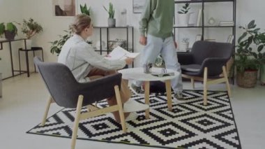 Genç bayan işaret dili öğretmeninin koltukta oturması, işitme engelli genç bir öğrencinin içeri girmesi, oturması, selamlaşması ve işaret diliyle iletişim kurması.