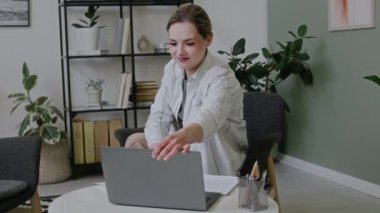 Genç beyaz kadının sınıfta ya da ofiste otururken, bilgisayarını açarken, video görüşmesi yapmak için tuşlarken, sonra online öğrenciyi selamlarken ve işaret dili hareketleriyle konuşurken orta eğik görüntüsü.