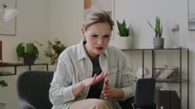 Orta halli, işitme engelli beyaz bir kadın tripodun üzerinde akıllı telefonun önünde oturmuş işaret dili kullanarak arkadaşlarıyla canlı bir sohbet ederken ve gülümserken.