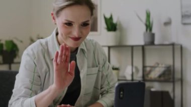 Sağır, dilsiz, beyaz bir kadının üç ayaklı telefondan video çağrısına cevap verirken, arkadaşına el sallarken, işaret dilini kullanırken, gülümserken ve gülerken orta eğik bir görüntü.