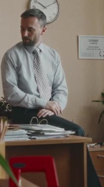 Beyaz tenli erkek katip memurun ekran dışından ahizeyi alıp iş meselelerini konuştuğu ve retro ofiste masa başında oturduğu dikey orta ölçekli bir fotoğraf.