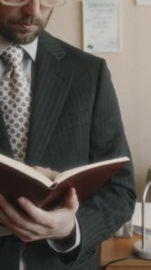 Kare çerçeveli gözlüklü, çizgili takım elbise ve kravat takan, not defterine not alan ve kameraya bakıp 90 'lı yıllarda modaya uygun bir ofiste poz veren sakallı beyaz bir patronun dikey eğik portresi.