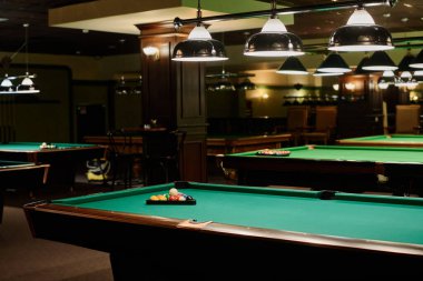 Modern bar ya da bardaki geniş salonun bir kısmında iki sıra yeşil bilardo masası ve üzerinde toplar olan üçgen raflar ve lambalar var.