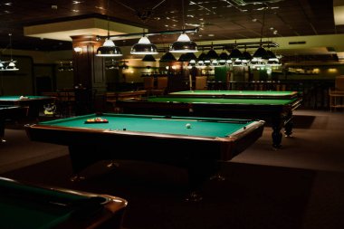 İki sıra ahşap bilardo masası, üzerinde yeşil tepeler ve lambaların bulunduğu geniş bir kulüp ya da bar salonu.