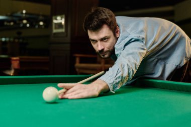 Açık mavi kot gömlekli genç adam bilardo oynarken bilardo masasına eğiliyor ve beyaz topa vurmaya konsantre oluyor.