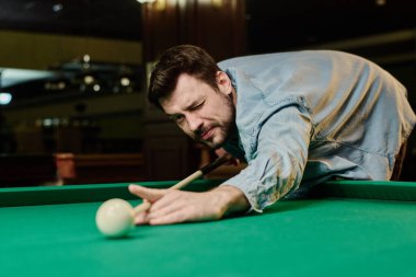 Genç adam bilardo masasına eğilip beyaz bilardo topuna bakarken sol gözünü kapalı tutuyor.