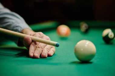 Hand of young man on top of green pool table holding billiard cue and concentrating on hitting white ball during play in game room clipart