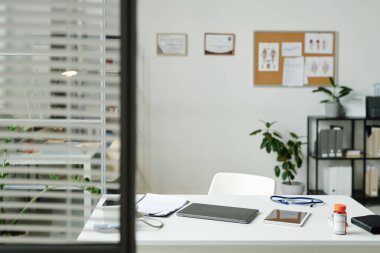 Modern pratisyen hekimin çalışma yerinde dizüstü bilgisayar, tablet, tıbbi belge, bir şişe hap, steteskop ve tonometre var.