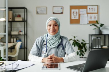 Tesettür ve tıbbi önlüklü genç İspanyol bayan tele-doktor iş yerinde dizüstü bilgisayarla otururken kameraya gülümseyerek bakıyor.