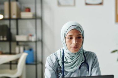 Genç Müslüman kadın sanal sağlık asistanı steteskobu ile çevrimiçi tıbbi danışmanlık sırasında dizüstü bilgisayara bakıyor.