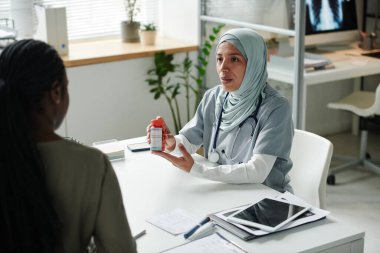 Genç, kendinden emin, tıbbi önlüklü, tesettürlü kadın klinisyen Afrikalı Amerikalı hastaya reçete yazan ve karakteristik özelliklerini tarif eden