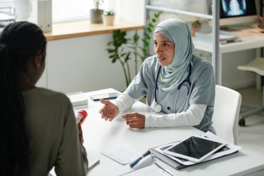 Tesettürlü ve tıbbi önlüklü genç İspanyol kadın danışmanlık sırasında bir şişe hap veya vitaminle kadın hastaya bakıyor.