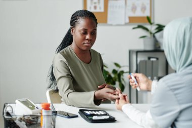 Genç hamile bir kadın klinikçinin önünde oturmuş muayene sırasında dijital tıbbi cihazla glikoz seviyesini kontrol ediyor.