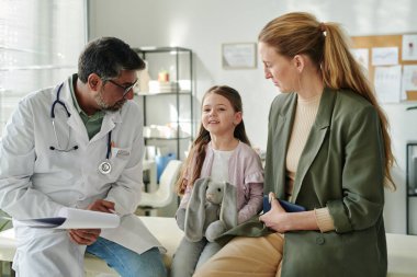 Annesi ve laboratuvar önlüğü giyen yetişkin erkek çocuk doktoru arasında oturan gri oyuncak tavşanı olan mutlu küçük kız.