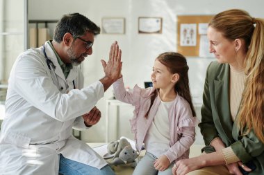Laboratuvar önlüklü ve gözlüklü mutlu çocuk doktoru annesinin yanında oturan yumuşak oyuncaklı şirin hastasına beşlik çakıyor.