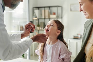 Ağzı açık küçük mutlu bir hastanın boğazını çelik spatula ile kontrol ettiriyorlar. Laboratuvar önlüğü giymiş yetişkin bir erkek çocuk doktoru tarafından tutuluyor.