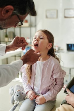 Çelik spatulası olan olgun erkek çocuk doktorunun elleri ağzını açık tutan küçük şirin bir hastanın boğazını kontrol ediyor.
