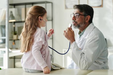 Olgun erkek çocuk doktoru, tıbbi danışmanlık yaparken steteskopla oynarken küçük şirin bir hastaya bakıyor.